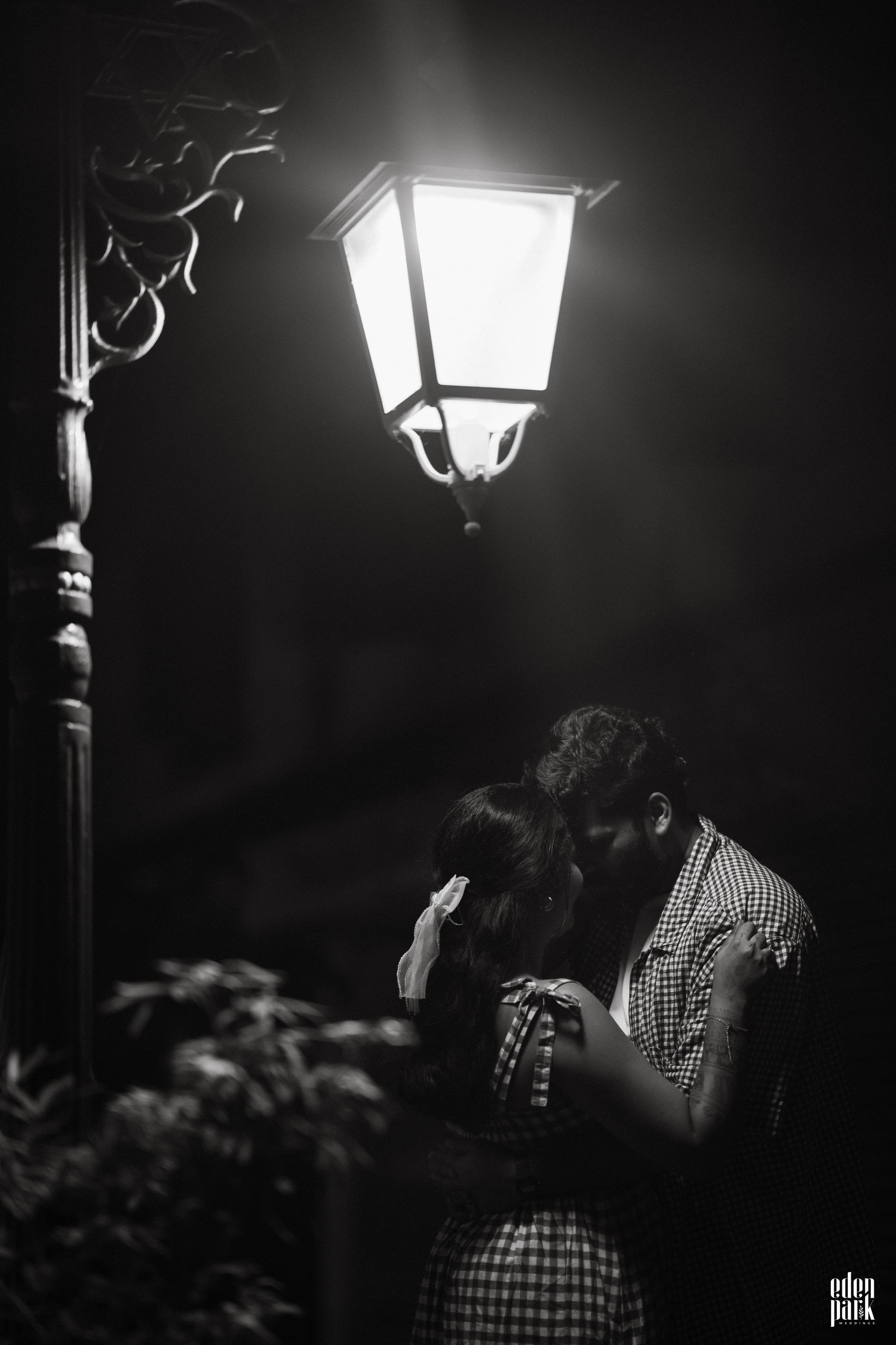 Black and white: a street lamp burning above a narrow lane at night, with Steeja and Arjun standing close together beneath it, foreheads almost touching.