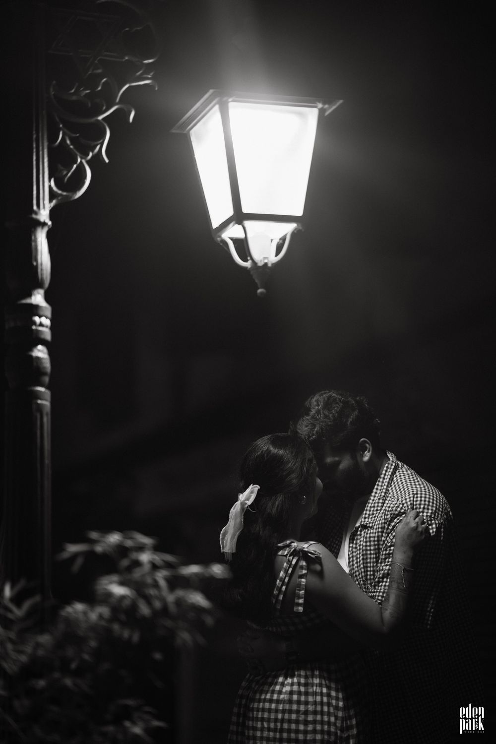 Black and white: a street lamp burning above a narrow lane at night, with Steeja and Arjun standing close together beneath it, foreheads almost touching.