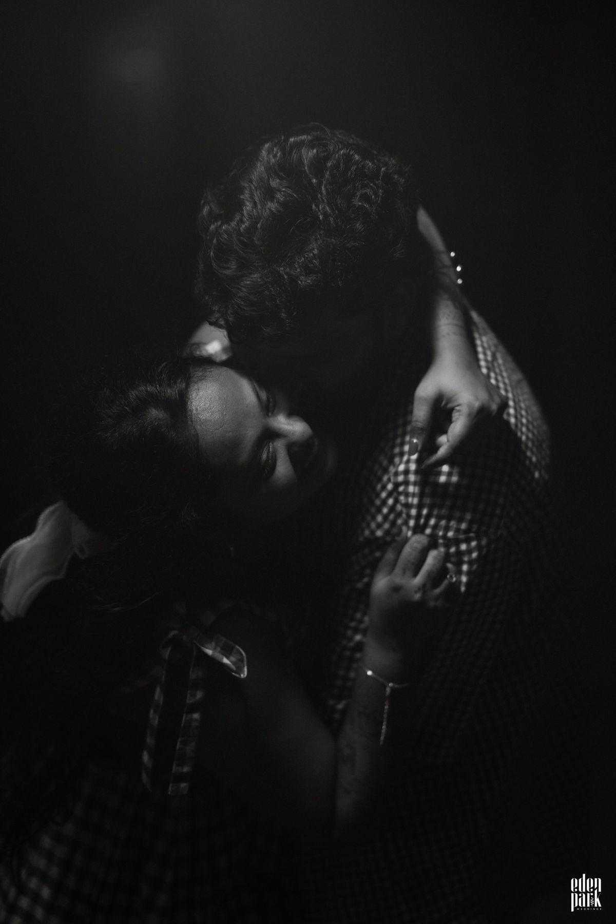 Black and white close-up of the two of them in the dark, her head tipped back mid-laugh against his shoulder, her hand resting on his chest.