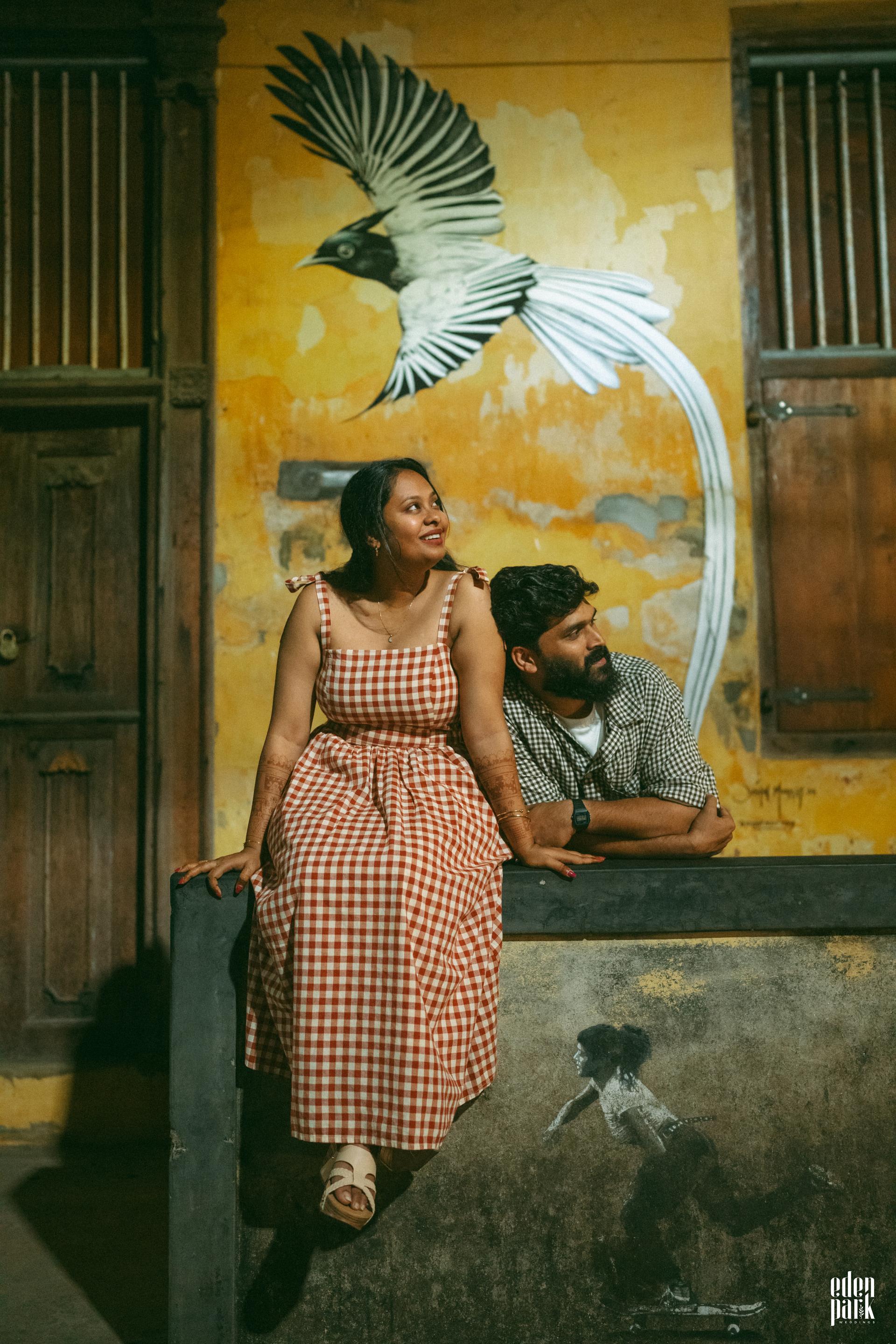 Steeja and Arjun sitting together on a ledge in front of a weathered ochre wall painted with a large white bird.