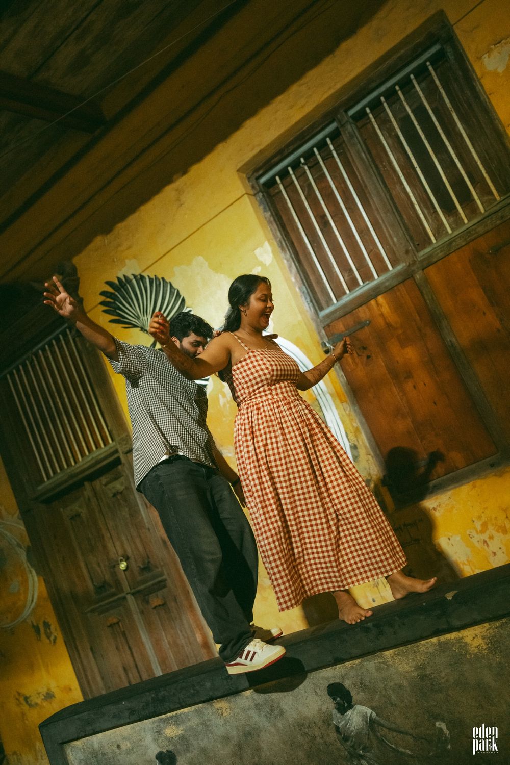 Steeja walking along a low ledge with her arms out for balance in front of the yellow wall, Arjun reaching up behind her.